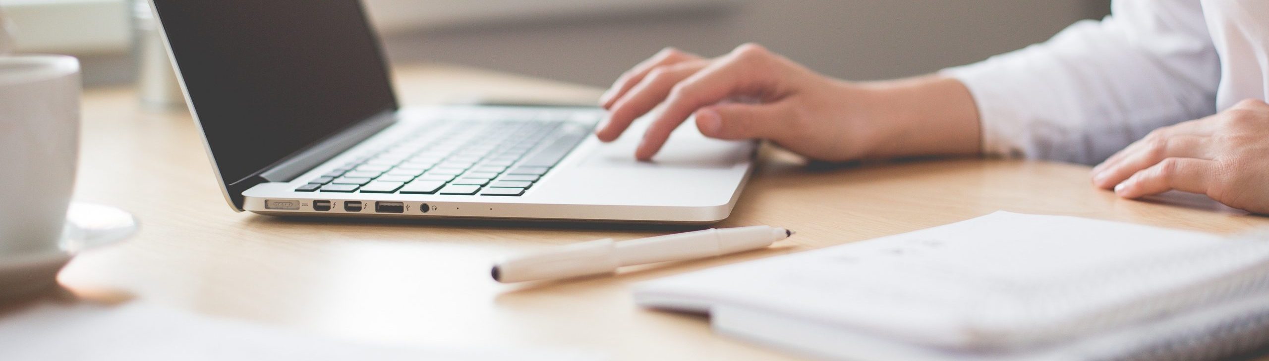 Een persoon is bezig achter de laptop. Er ligt een schrift met een pen op tafel