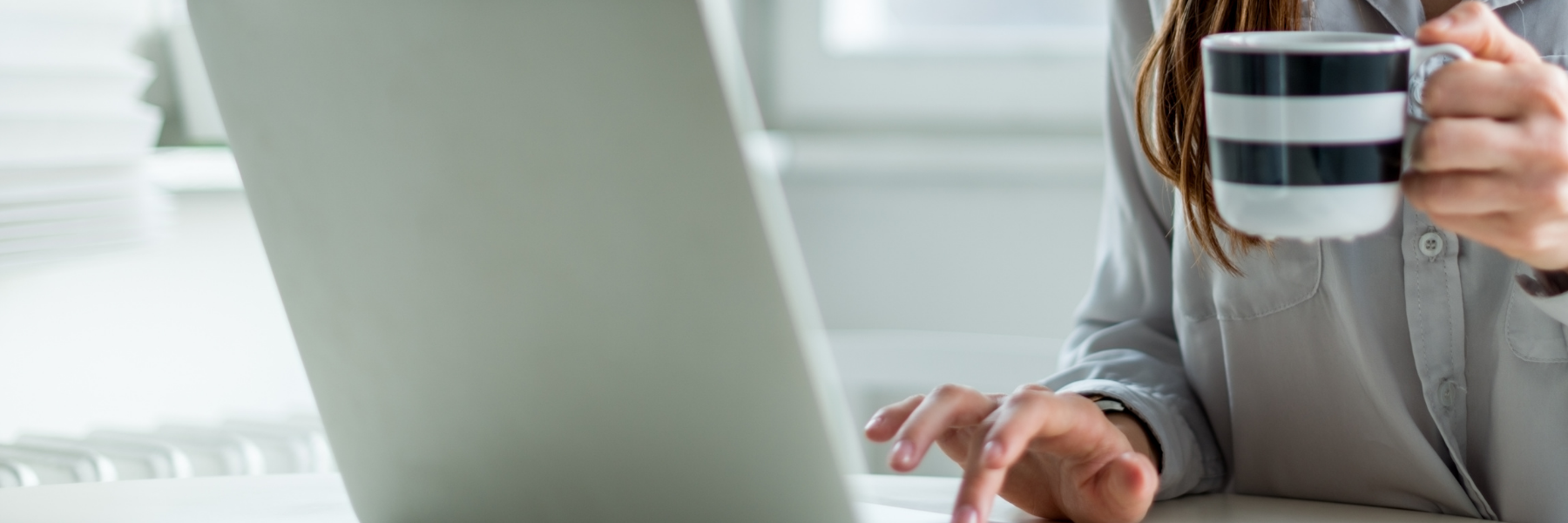 Een persoon zit achter de laptop met één had te typen en in de andere hand een mok met koffie